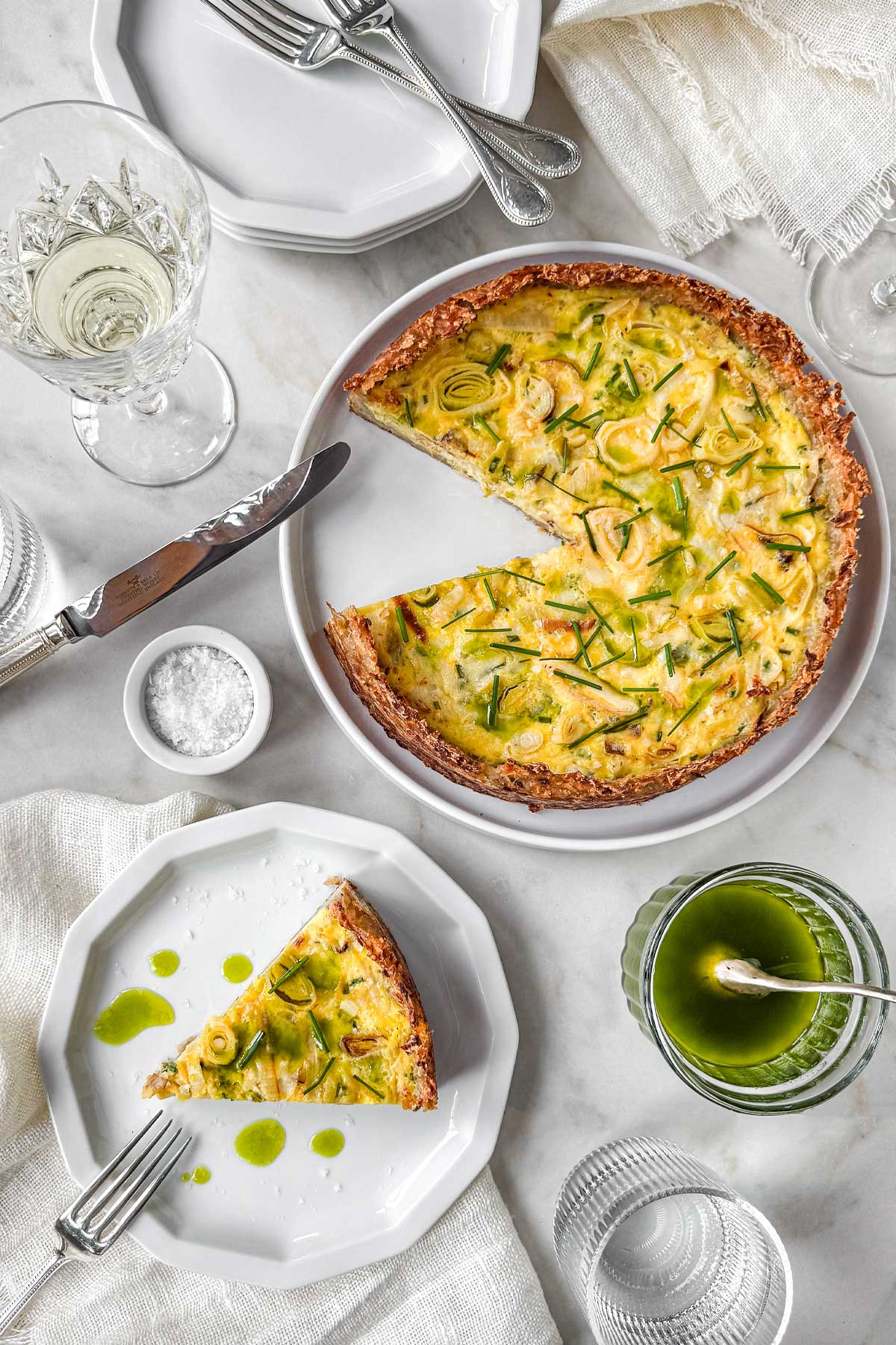 Hashbrown crust quiche with leeks and Gruyère topped with chives and drizzled chive oil, served on a marble table with a sliced piece on a plate