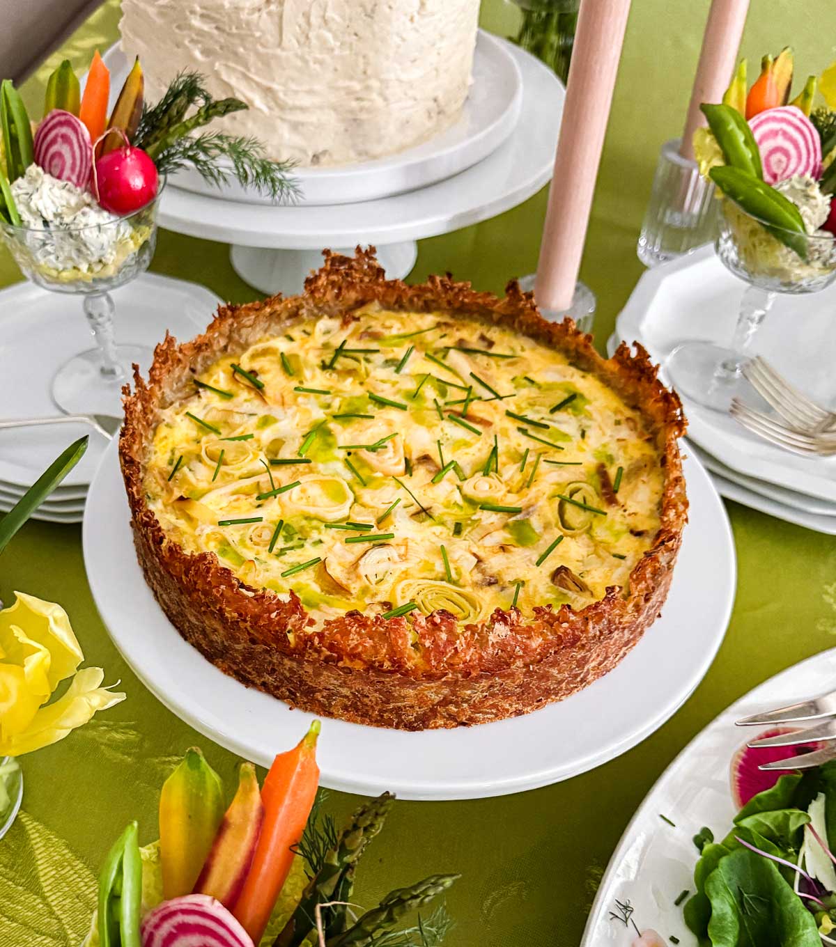 A Hashbrown Crust Quiche on a spring brunch table on a white cake stand surrounded by spring appetizers.
