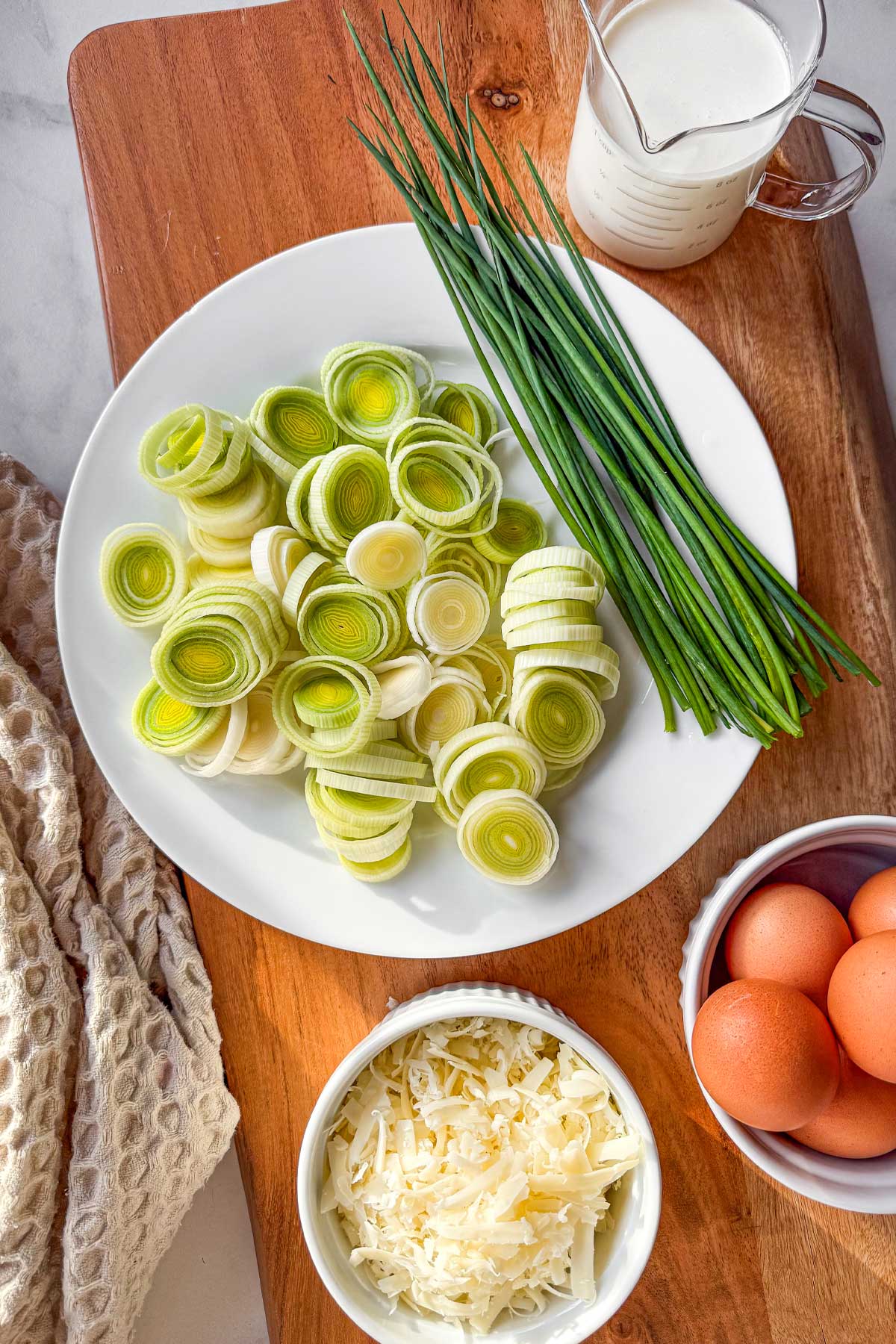 The ingredients for hashbrown crust quiche with leeks, gruyere, and chives in bowls on a wooden board.