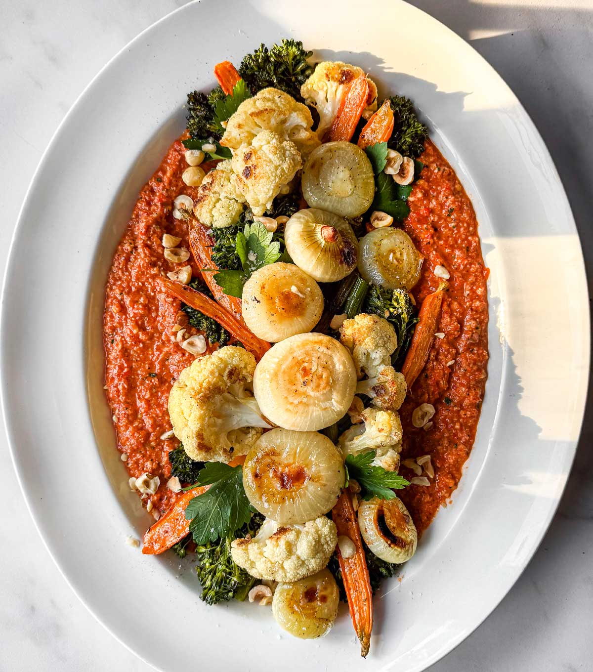 A white oval serving platter with warm orange Romesco sauce layered with roasted winter vegetables: cippolini onions, broccolini, carrots, and cauliflower, finished with crushed hazelnuts.