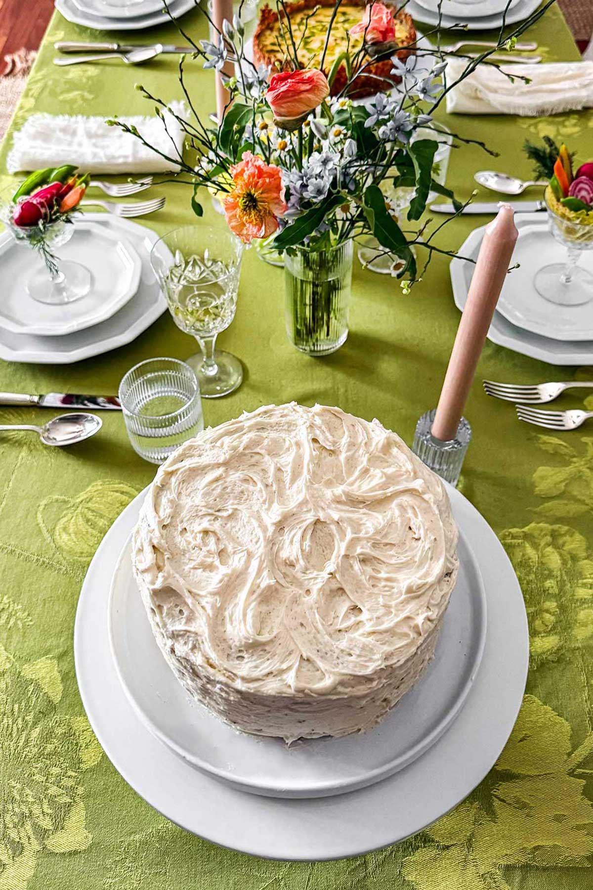 Spring brunch table with green linen, fresh floral centerpiece, garden crudité goblets, layered white plates, and a carrot cake for Easter or Mother’s Day entertaining