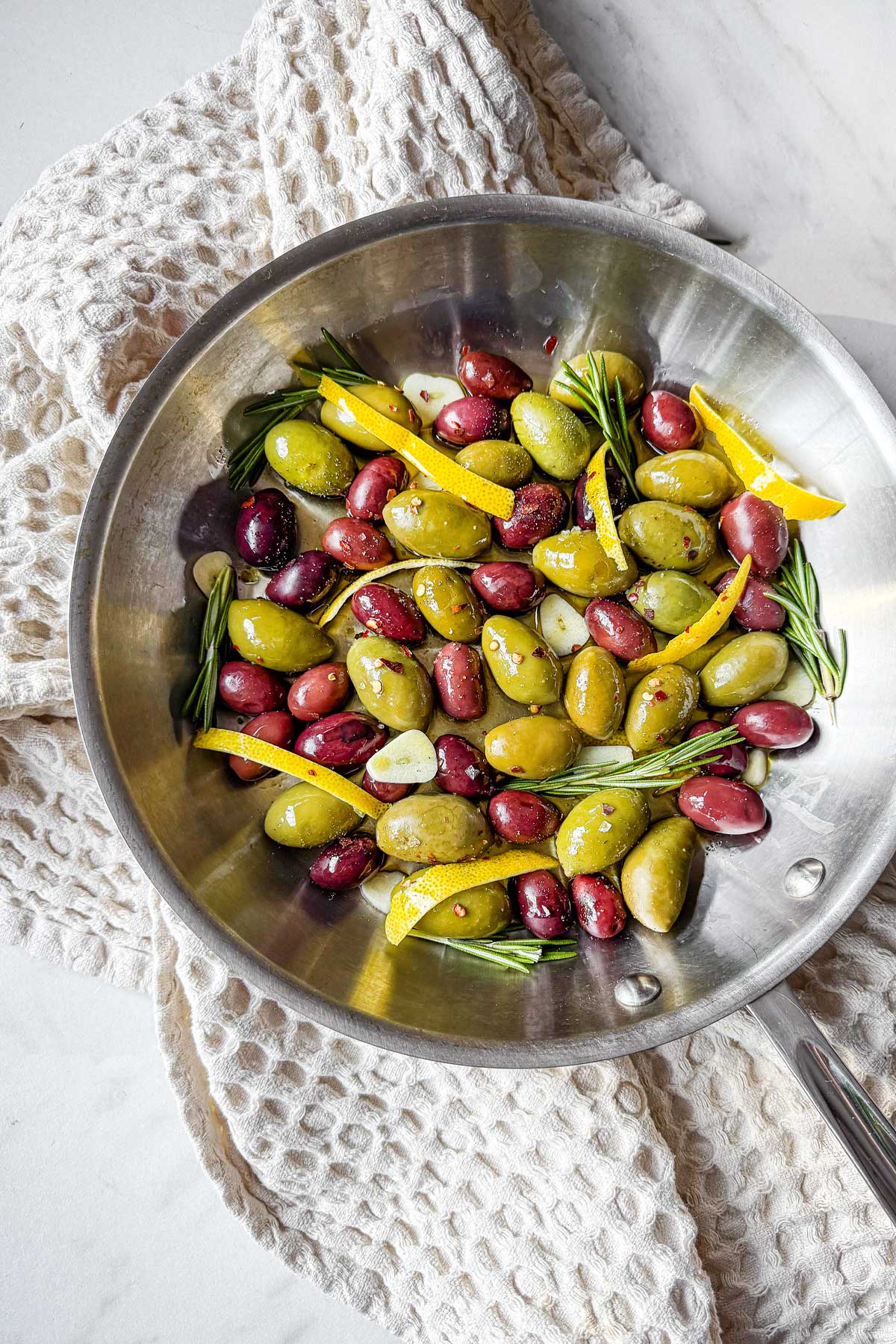 A skillet filled with mixed olives, citrus peel, rosemary sprigs, olive oil, and spices, sitting on a waffle tea towel.