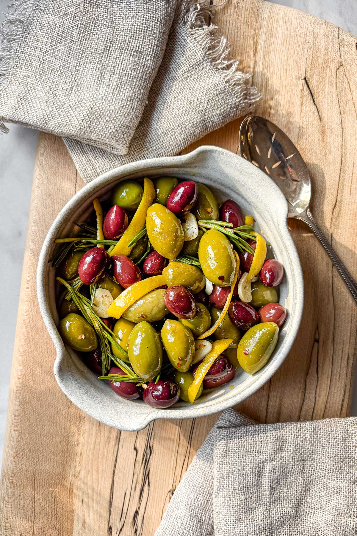 An artisanal ceramic bowl filled with warm citrus marinated olives for snacking, with linen cocktail napkins for serving.