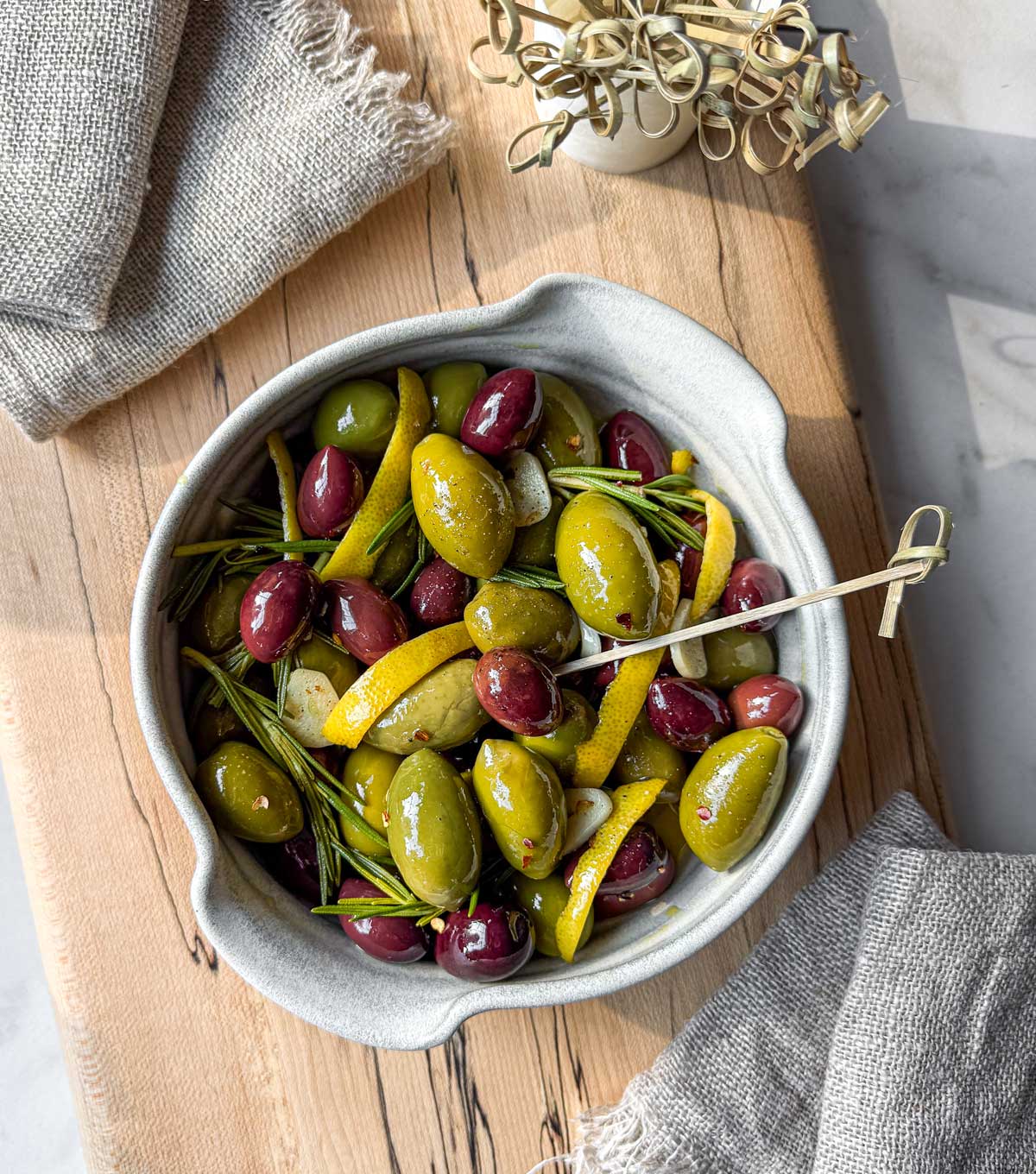 An artisanal ceramic bowl filled with warm citrus marinated olives for snacking, with bamboo cocktail picks and linen cocktail napkins for serving.