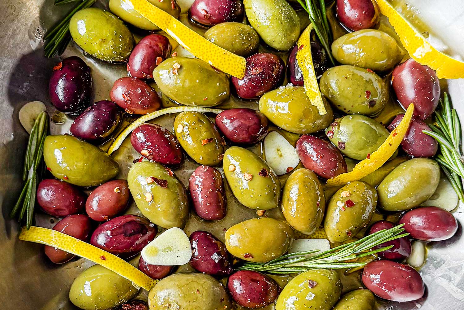 A skillet filled with mixed olives, citrus peel, rosemary sprigs, olive oil, and spices, sitting on a waffle tea towel.
