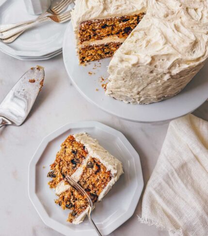 A slice of carrot cake with brown butter cream cheese frosting on a plate next to a layered carrot cake on a white pedastel.
