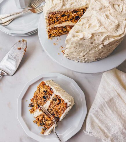 A slice of carrot cake with brown butter cream cheese frosting on a plate next to a layered carrot cake on a white pedastel.