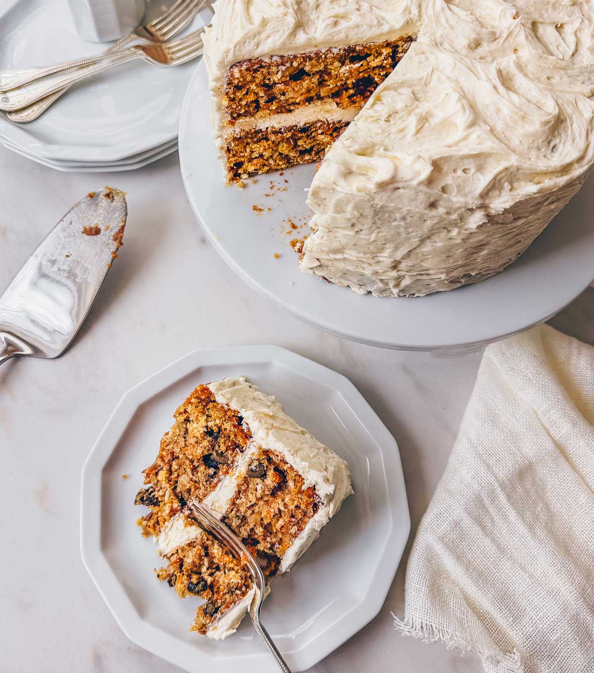 A slice of carrot cake with brown butter cream cheese frosting on a plate next to a layered carrot cake on a white pedastel.